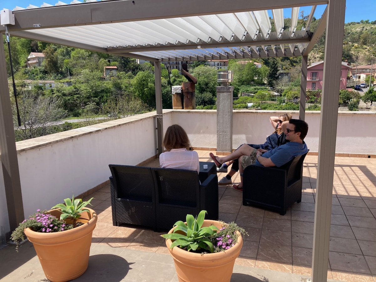 A spacious terrace is featured, equipped with a shaded pergola. Two comfortable chairs with cushions are positioned next to potted plants. The scenic countryside is visible in the background, showcasing greenery and traditional homes, enhancing the outdoor relaxation space.
