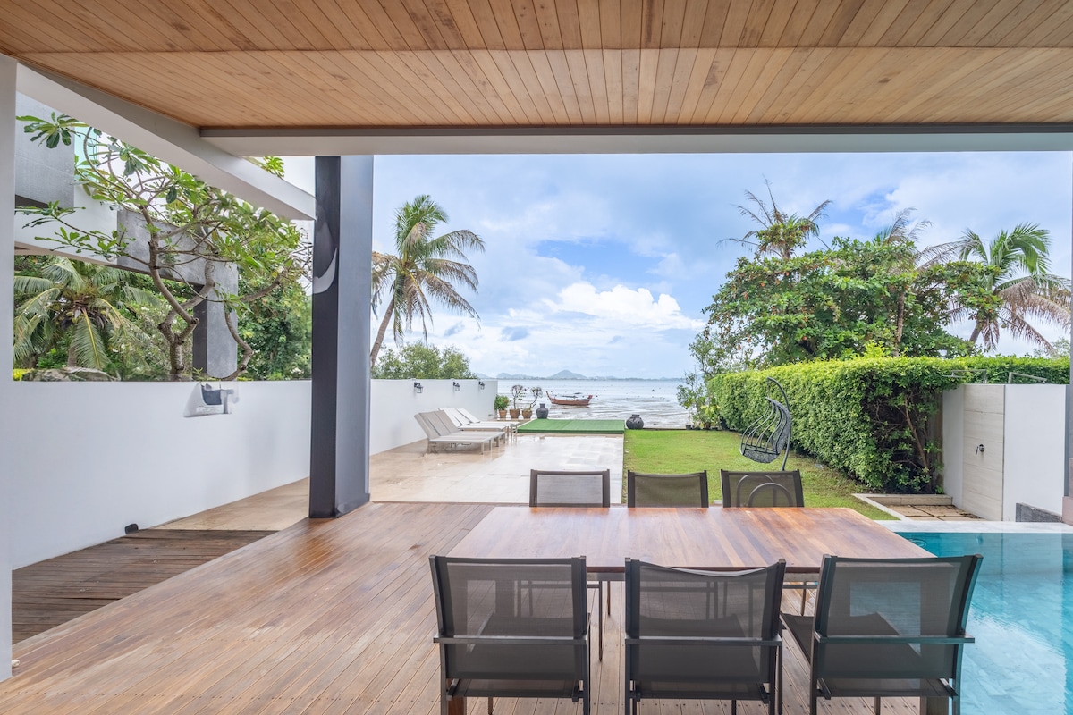 An outdoor patio area is featured, showcasing a wooden dining table surrounded by black chairs. A swimming pool glistens nearby, with a well-maintained garden and palm trees visible in the background. The scenic view of the ocean is presented through large open spaces.