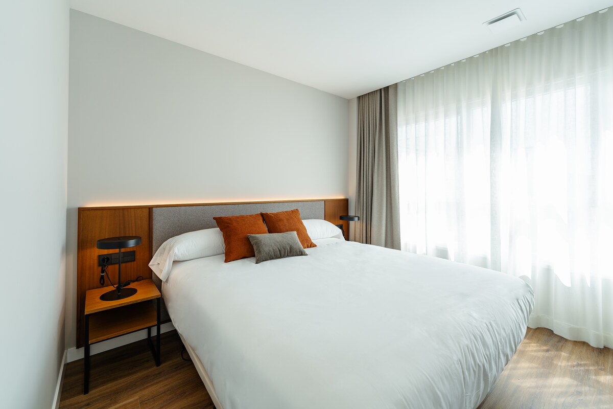 A minimalist bedroom features a spacious bed with neutral bedding and accent pillows. Stylish bedside tables with lamps are positioned on either side. Soft natural light filters through sheer curtains, highlighting the light wood floors and modern decor.