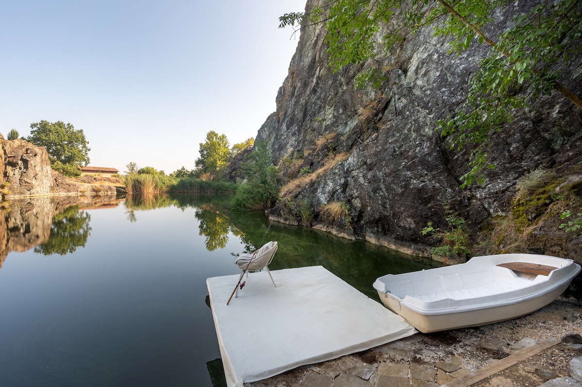 A serene lakeside area is highlighted, featuring a smooth, white platform with a single chair positioned for relaxation. A small boat rests nearby on the water's edge, while the calm lake reflects surrounding trees and rocky cliffs.