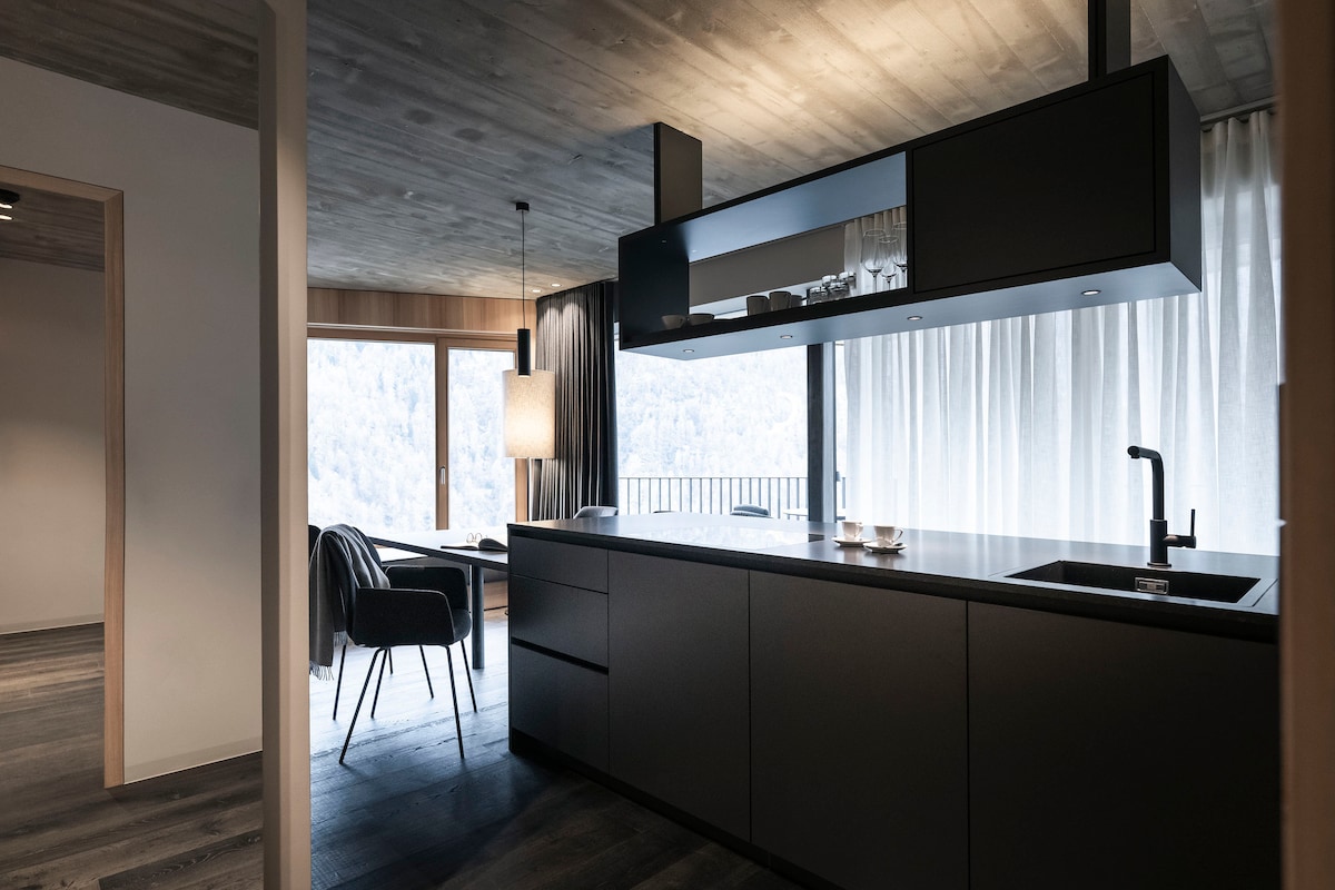 A modern kitchen features sleek black cabinetry and a spacious countertop. Natural light filters through large windows, illuminating the area. A dining table with chairs sits nearby, while a cozy armchair adds comfort to the open layout.