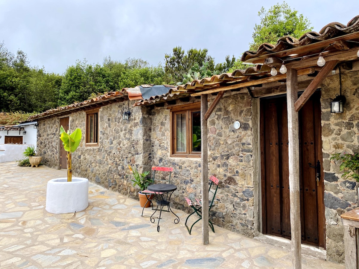 A rustic stone exterior is complemented by a tiled terrace and wooden beams. A small round table with two chairs is placed near an agave plant. The entrance features wooden double doors framed by soft outdoor lighting and surrounded by lush greenery.