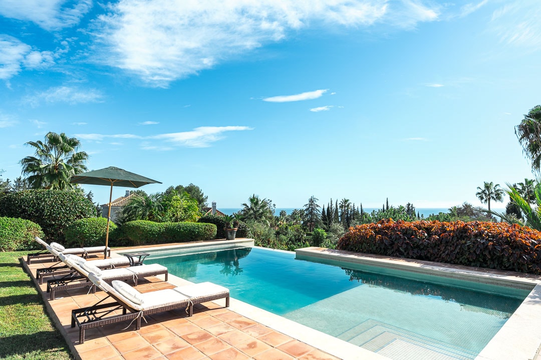 An inviting view of the private infinity pool surrounded by lush gardens and palm trees. The pool features ten sun loungers arranged neatly along its edge, with a green parasol providing shade. A clear blue sky complements the serene atmosphere.