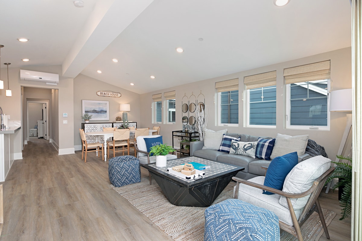 A spacious living area features comfortable furniture arranged for gatherings. Natural light enters through multiple windows with stylish shades. A dining table for six is visible in the background, alongside a cozy seating area adorned with decorative pillows and a coffee table displaying various items.