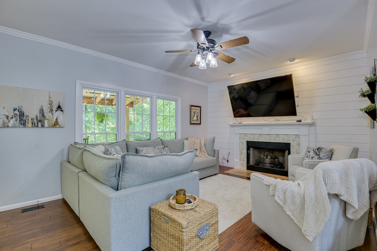 A bright living area features ample seating with light gray sofas arranged around a cozy fireplace. Natural light streams in through multiple windows, enhancing the space. A flat-screen television is mounted above the fireplace, and a textured rug complements the wooden flooring.