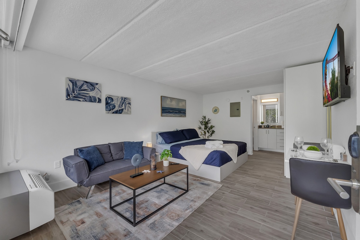 A modern studio space features a cozy seating area with a dark sofa beside a small coffee table. A spacious bed with blue linens is positioned against the wall. A flat-screen TV is mounted on a light wall, and a kitchen area is visible in the background.