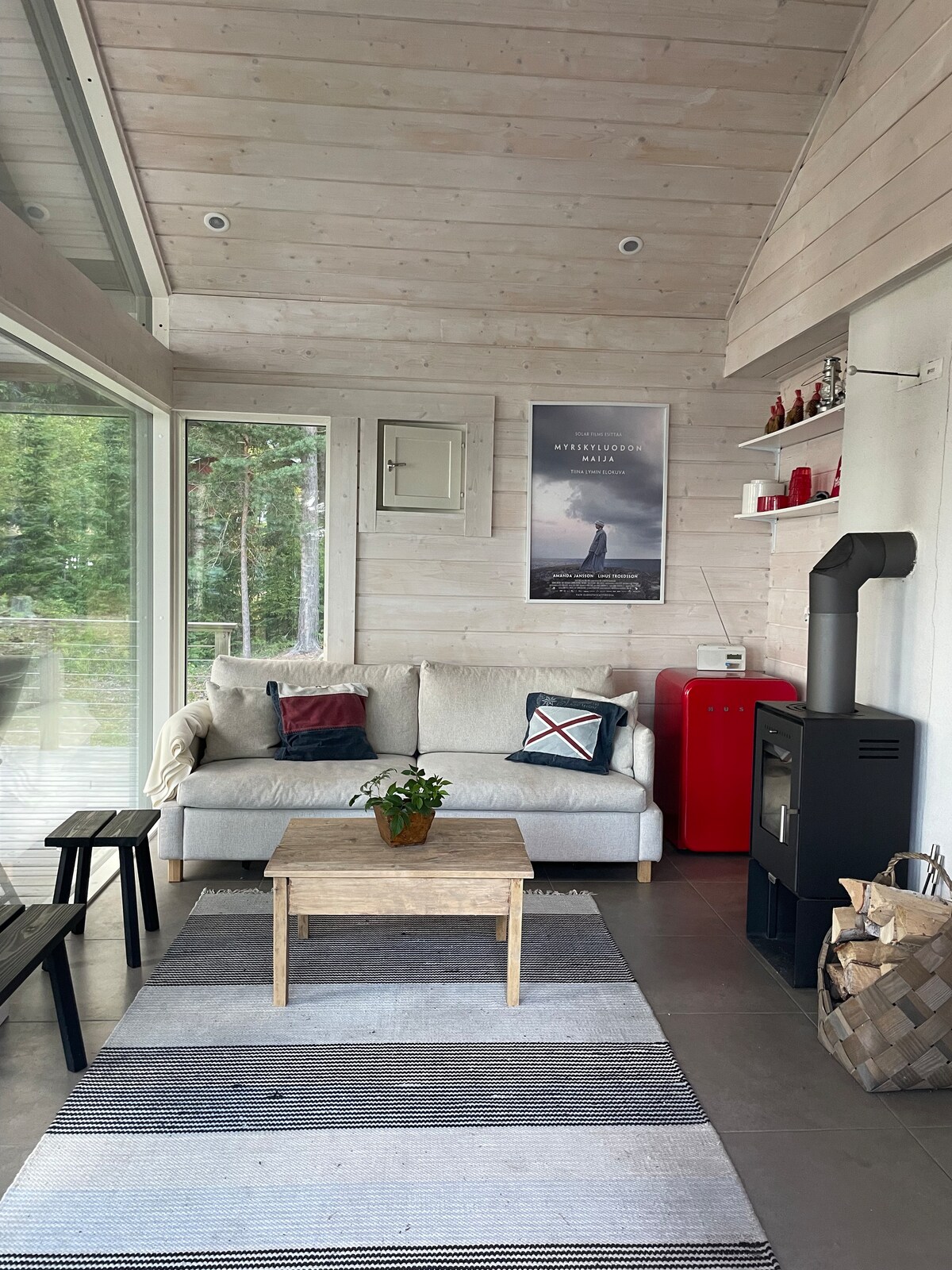 An inviting living area features a comfortable sofa adorned with decorative pillows. A small wooden coffee table sits on a striped area rug. A sleek red refrigerator adds a pop of color, while a modern stove provides warmth against the backdrop of large windows overlooking nature.