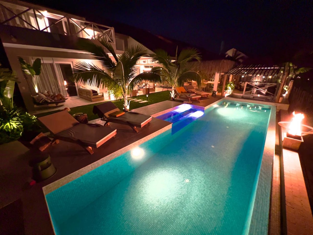 An illuminated pool area is surrounded by lush palm trees and cozy lounge chairs. The clear water glows invitingly, complemented by warm outdoor lighting. A spacious terrace and tropical landscaping enhance the atmosphere, creating a serene outdoor retreat suitable for relaxation and leisure.