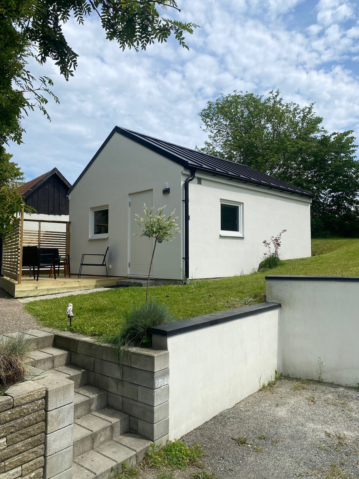 A small, white cottage is set on a grassy area, framed by trees. A concrete pathway leads to the front entrance, which features a single door. The structure is accompanied by a sunny outdoor seating area with chairs visible in the background.