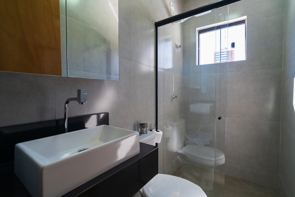 A modern bathroom features a sleek countertop with a rectangular sink. A glass shower enclosure is visible, highlighting grey tiles and contemporary fixtures. Natural light is provided by a small window, and a toilet is positioned discreetly in the corner.