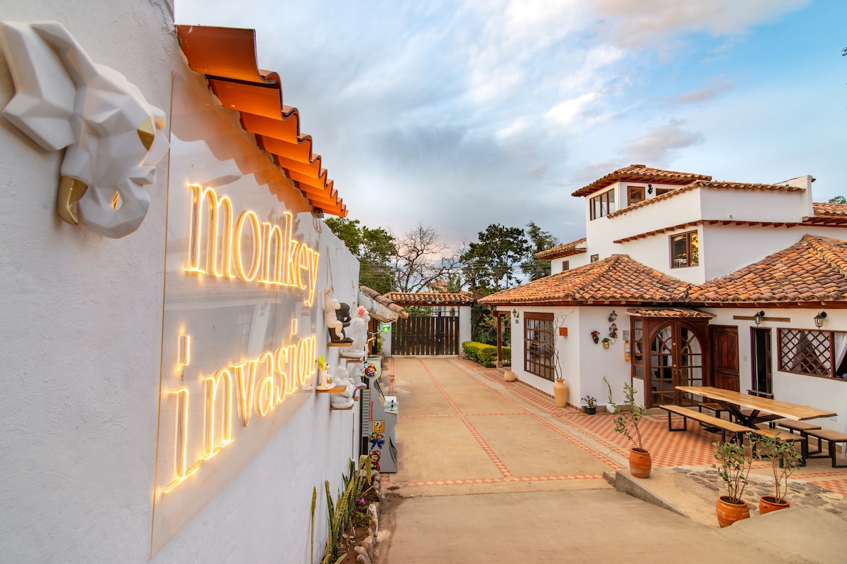 A charming entrance is showcased with a decorative sign reading 'Monkey Invasion.' The pathway is flanked by plants, leading to a welcoming two-story building with a tiled roof. Large windows and a spacious outdoor dining area are visible, enhancing the inviting environment.
