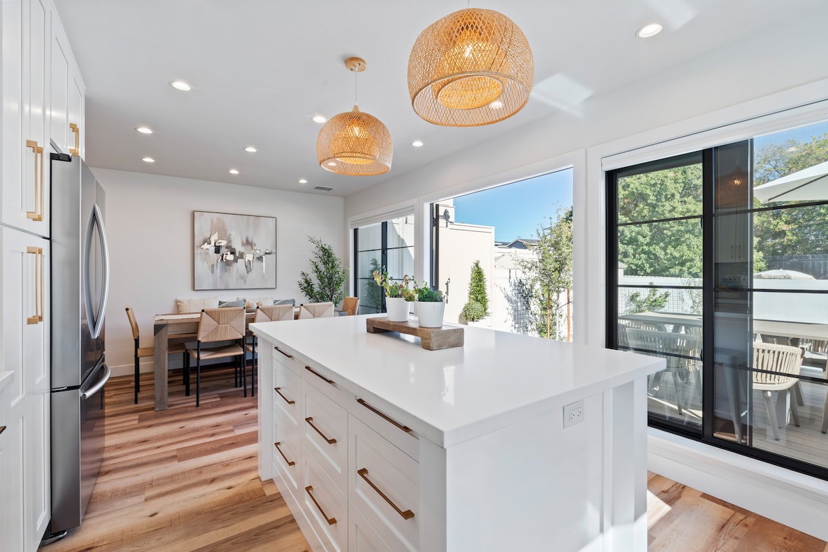 A modern kitchen showcases an expansive white island with sleek cabinetry and stainless steel appliances. Two woven pendant lights hang above, illuminating the space. Large glass doors provide views of the outdoor patio and dining area, enhancing the inviting atmosphere.