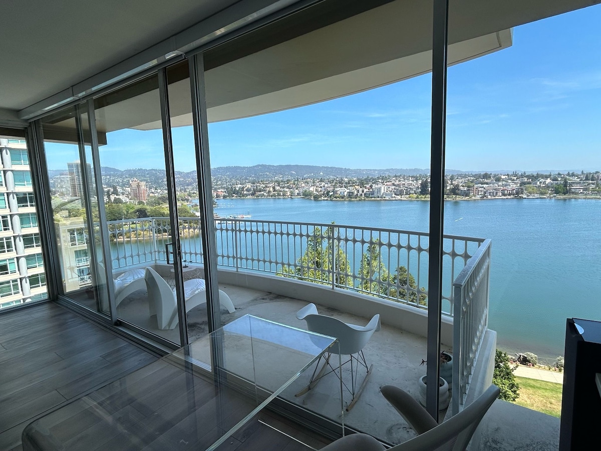 The image captures a spacious living area with large glass doors that open to a balcony overlooking Lake Merritt. A transparent dining table is visible, complemented by modern, white chairs. Sunlight floods the room, highlighting the serene blue water and distant city views.