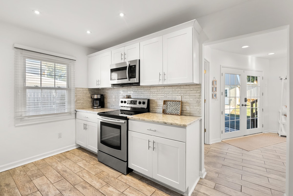 The kitchen is equipped with modern appliances, including a stove, microwave, and coffee maker. White cabinets and a textured backsplash complement the light countertops. Natural light fills the space through a nearby window, while tile flooring adds a practical touch.