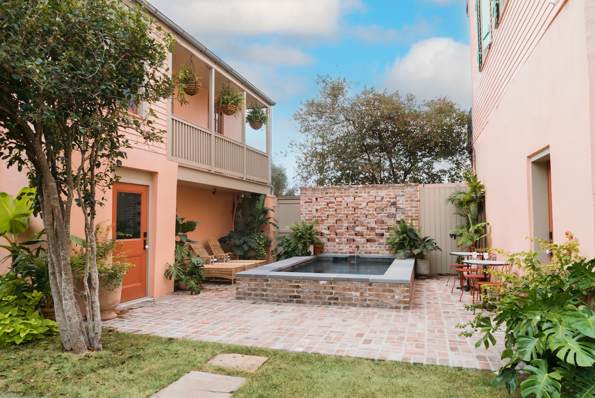 A serene courtyard is depicted, featuring a plunge pool surrounded by brick and lush greenery. The light-colored walls of the surrounding buildings create a welcoming atmosphere. Comfortable chaise lounges are situated nearby, inviting relaxation in this tranquil outdoor space.