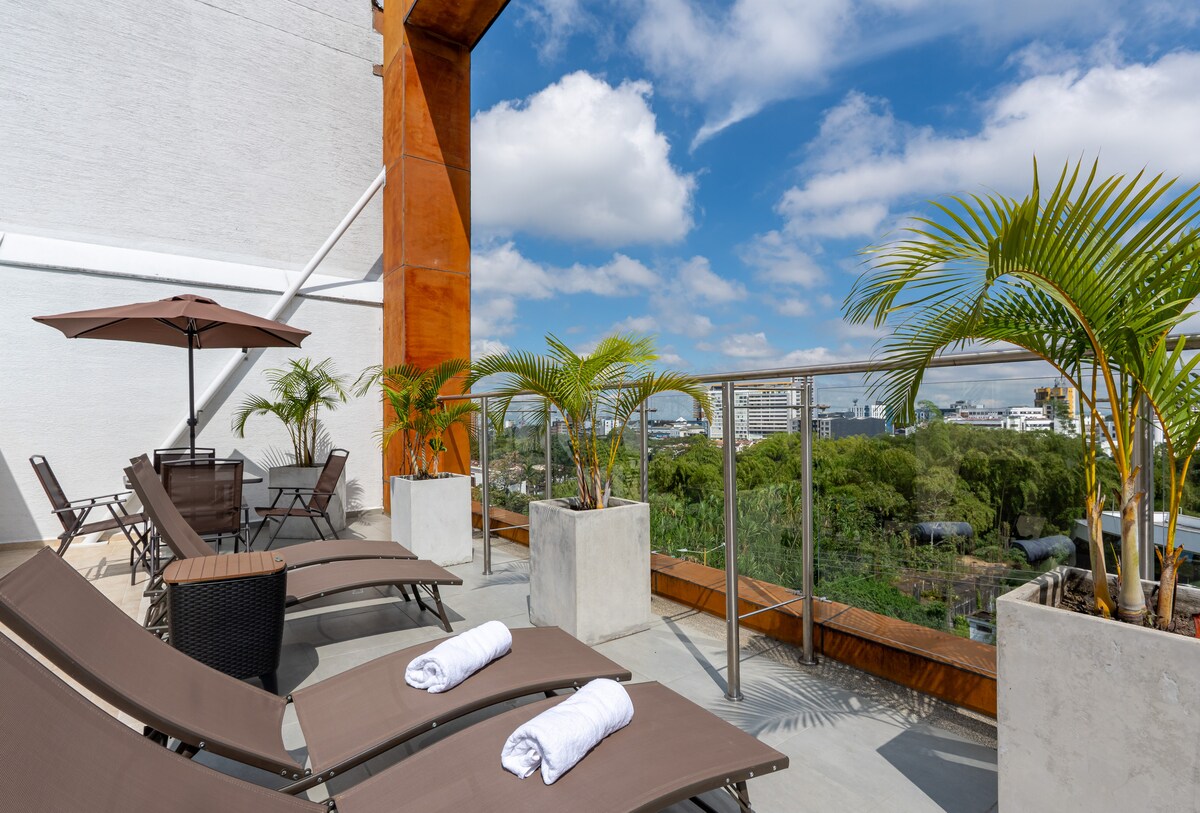 A rooftop terrace features lounge chairs positioned under a shade umbrella, offering a view of the surrounding landscape. Potted palm trees are present, and neatly rolled towels are placed on each chair. The sky is bright with scattered clouds, enhancing the outdoor experience.