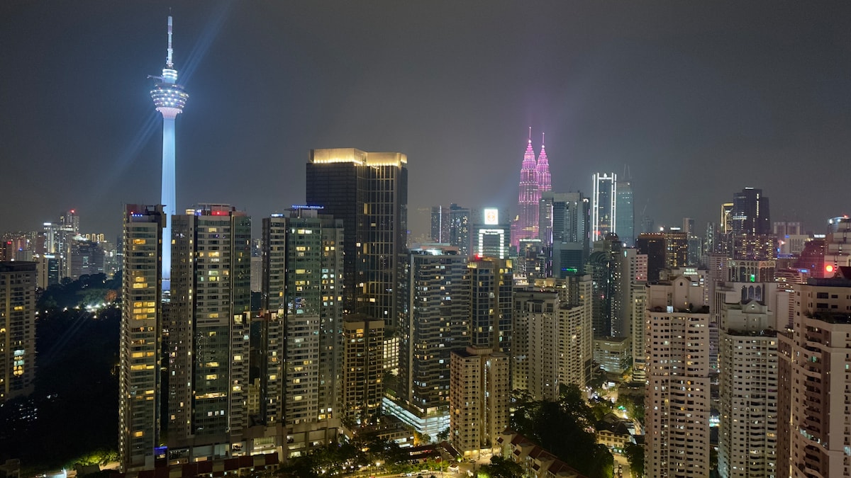 A panoramic view of the city skyline at night, featuring illuminated buildings and the iconic Twin Towers and KL Tower. A vibrant display of lights creates a lively urban atmosphere, showcasing the modern architecture and lively surroundings.