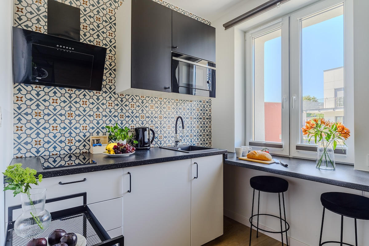 The kitchen area features a modern design with patterned tiles on the backsplash. A sleek countertop is complemented by essential appliances, including a microwave and kettle. A small dining nook with two stools is situated beside a window, offering natural light and a view.