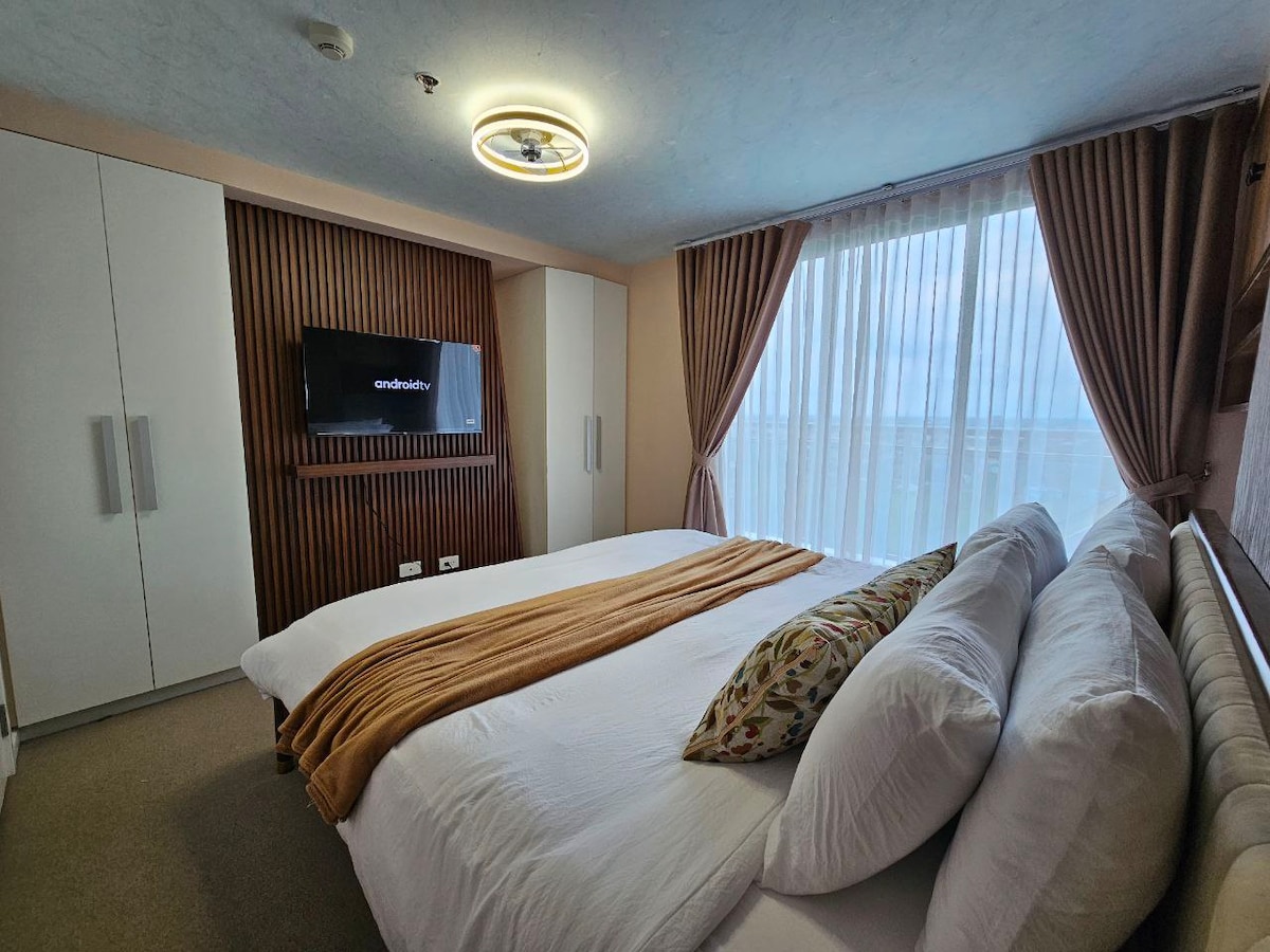 A cozy bedroom features a spacious bed adorned with pillows and a decorative throw. A wall-mounted TV is positioned above a cabinet, and large windows dressed with sheer curtains allow natural light to fill the room.