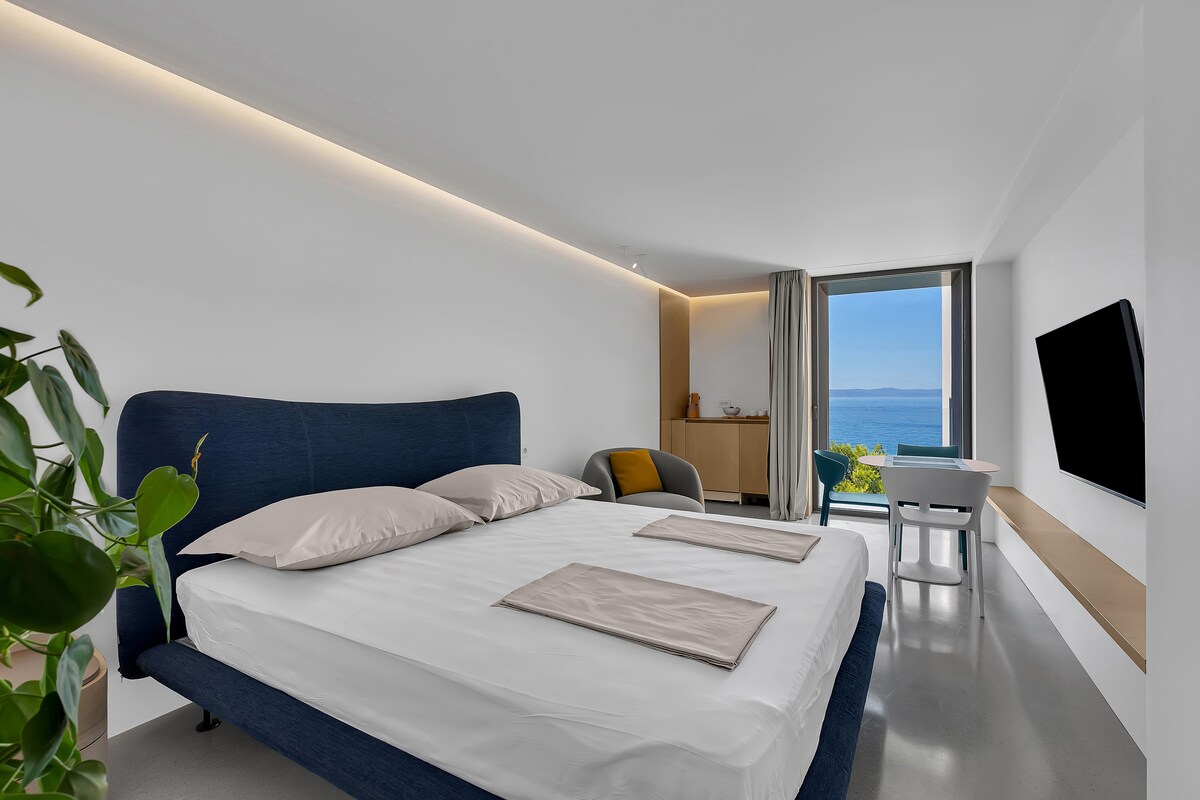 A bright bedroom features a spacious bed with neutral-colored linens and two pillows. A wall-mounted TV is positioned above a minimalist console. Natural light fills the room through a large window that offers views of the ocean. A potted plant adds a touch of greenery.