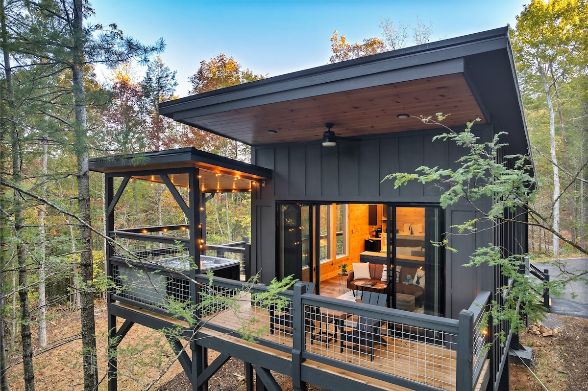 The exterior of the modern treehouse features a wraparound deck surrounded by trees, with soft lighting outlining the edges. Large glass doors provide a view into the inviting interior, while the roof extends over both the deck and living area, creating a seamless connection with nature.