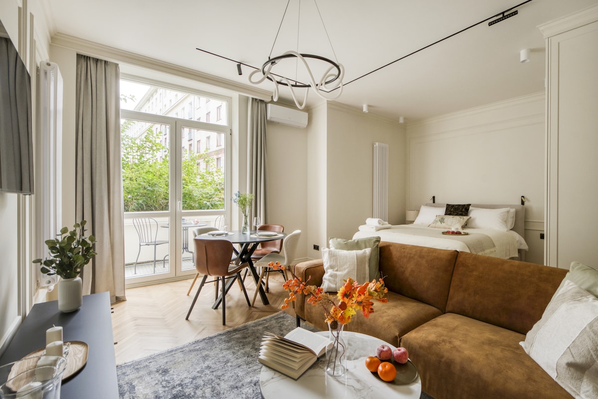 A bright living space features a comfortable brown sofa and a round dining table with stylish chairs. Sunlight filters through large glass doors, which provide access to a private balcony overlooking greenery. Decorative elements include a vase with flowers and fruits on the table.