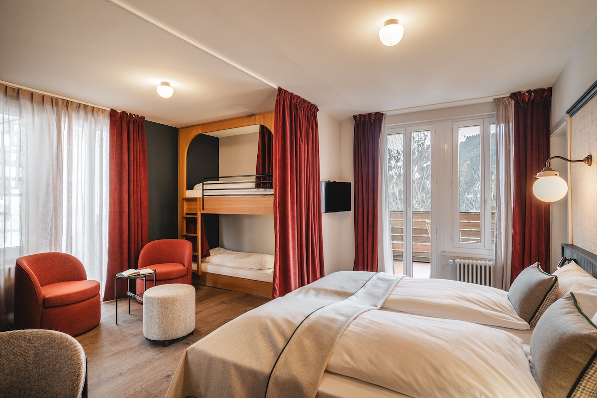 A spacious guest room features two beds positioned side by side, complemented by a cozy bunk bed in the background. Natural light filters through large windows adorned with curtains. A small round ottoman and two armchairs are placed near a low table, enhancing the inviting layout.