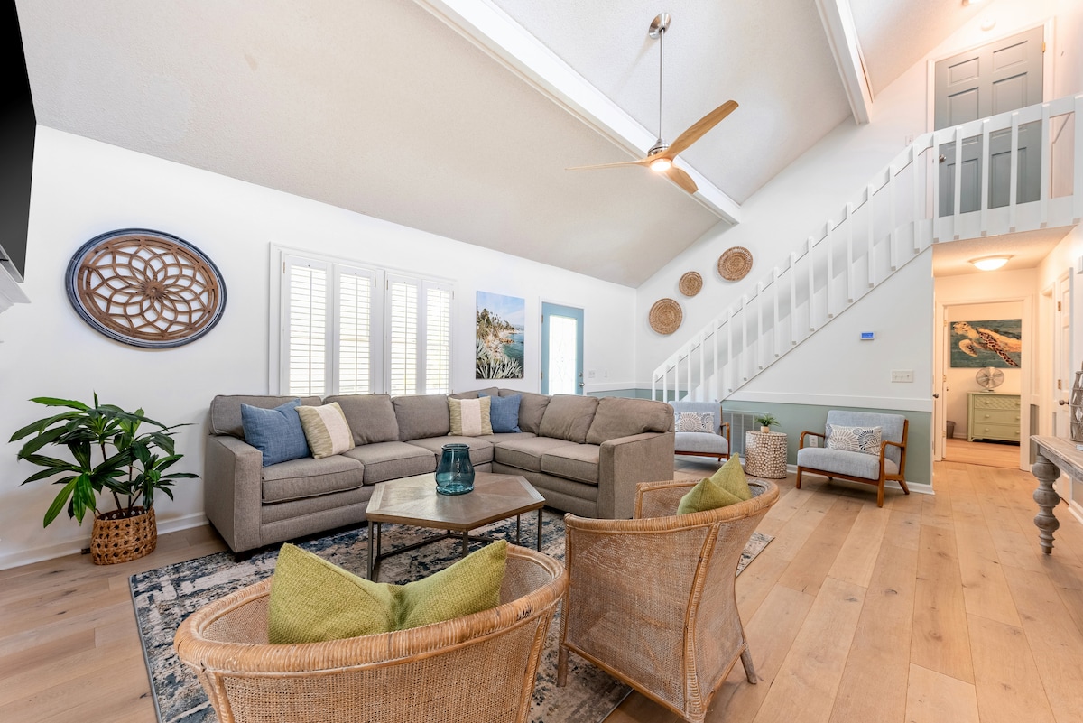 A spacious living area features a sectional sofa with decorative cushions, paired with wicker armchairs surrounding a wooden coffee table. Large windows with plantation shutters allow natural light to fill the room. A staircase leads to the upper level, accentuated by unique wall decor.