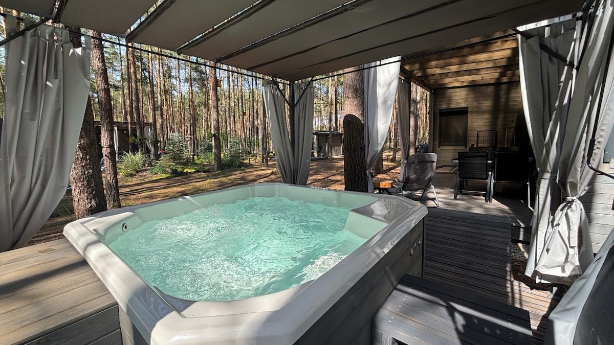 A private jacuzzi is enclosed under a shaded canopy, surrounded by tranquil pine trees. The bubbling water creates a relaxed atmosphere, while a cozy seating area with two chairs is positioned nearby, providing a space for guests to unwind in a natural setting.