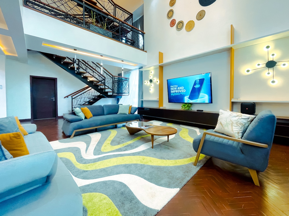 The spacious living area showcases modern furniture arranged around a vibrant area rug featuring a wavy design in shades of green and beige. A large flat-screen TV is mounted on a dark cabinet against the wall, with bright natural light filtering in from large windows.