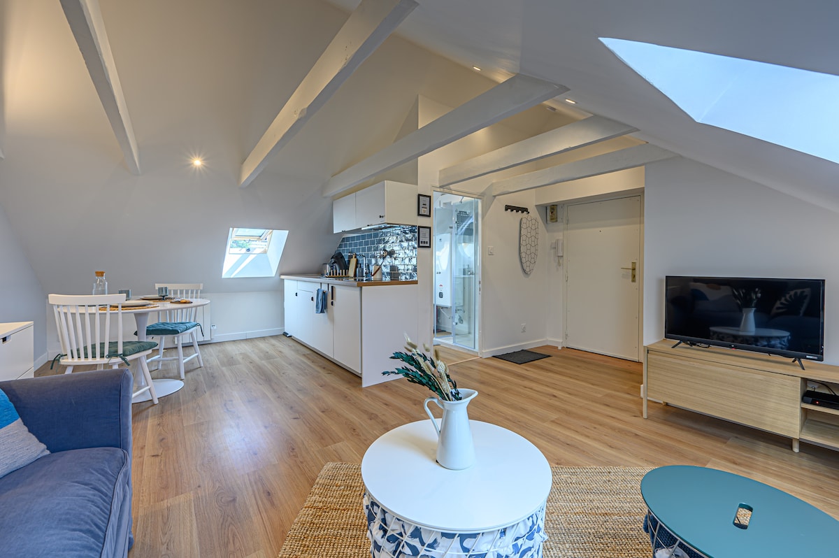 The studio showcases an open layout with wooden floors and slanted ceilings. Natural light floods the space through skylights. A cozy seating area is paired with a simple coffee table, while a dining area features two chairs and a small table. The kitchenette is visible in the background.