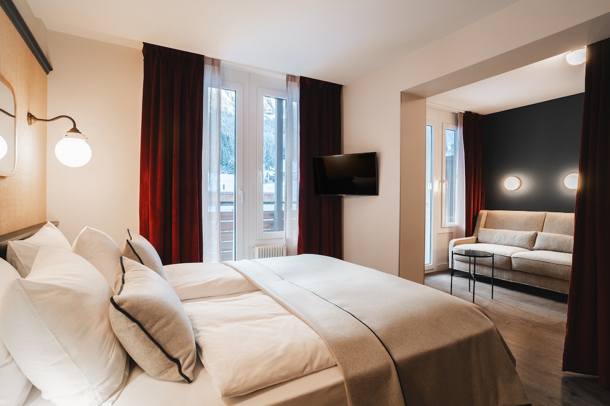 A modern guest room features a spacious bed with crisp white linens and a cozy beige throw. A comfortable sofa is positioned in a separate seating area, while large windows with sheer curtains allow natural light to fill the room.