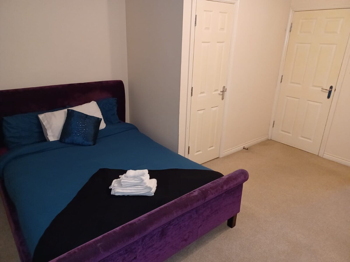 A spacious bedroom features a plush purple bed frame with a neatly made bed adorned in blue linens. Two doors provide access to other areas, while a stack of fresh towels rests on the bed, enhancing the inviting nature of the room.