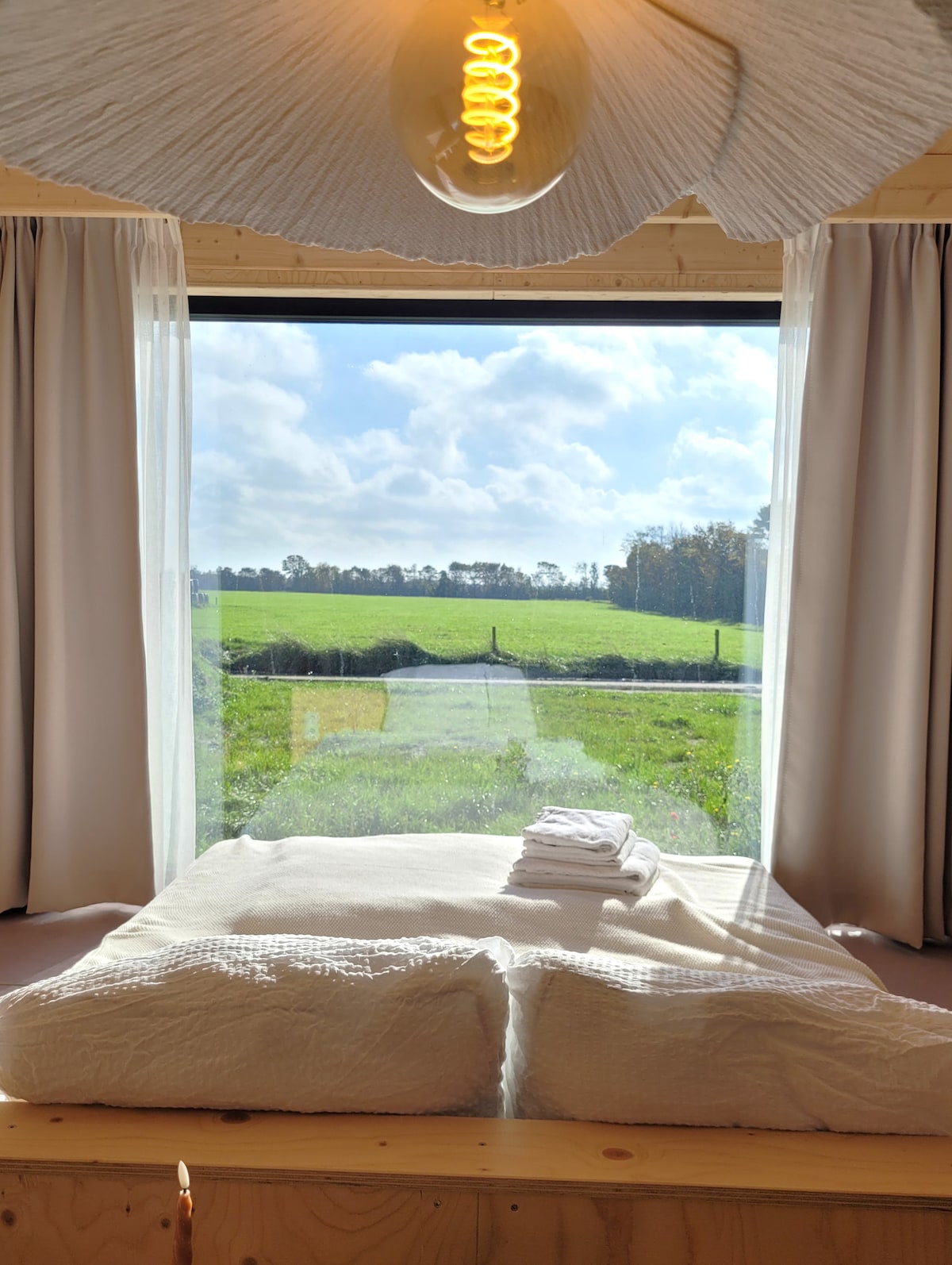 A spacious bed with white linens is positioned in front of large windows. Sunlight fills the room, illuminating scenic views of green fields and blue skies. Soft curtains frame the window, enhancing the connection to the serene outdoor landscape.