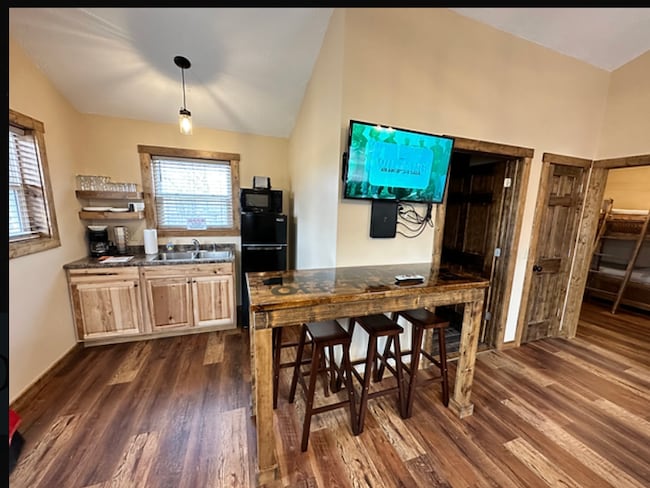 A kitchenette area features wooden cabinetry, a countertop with three bar stools, and a flat-screen TV mounted on the wall. A refrigerator and sink are visible, with natural light filtering through a window. The flooring consists of warm, wooden planks.