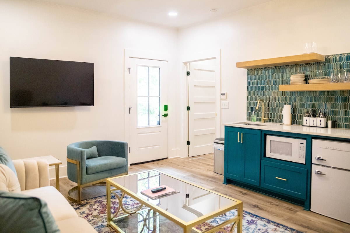 The kitchenette features a vibrant turquoise cabinetry with a microwave, mini-fridge, and coffee maker. A sleek flat-screen TV is mounted on the wall. A comfortable chair offers seating near a glass coffee table atop a decorative rug. Natural light enters through the door and nearby window.
