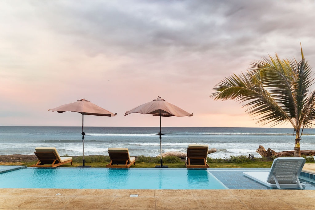 A private pool is shown with three lounge chairs underneath large umbrellas, facing an expansive ocean view. Gentle waves can be seen in the distance, with a palm tree partially visible on the right, set against a softly colored sky.