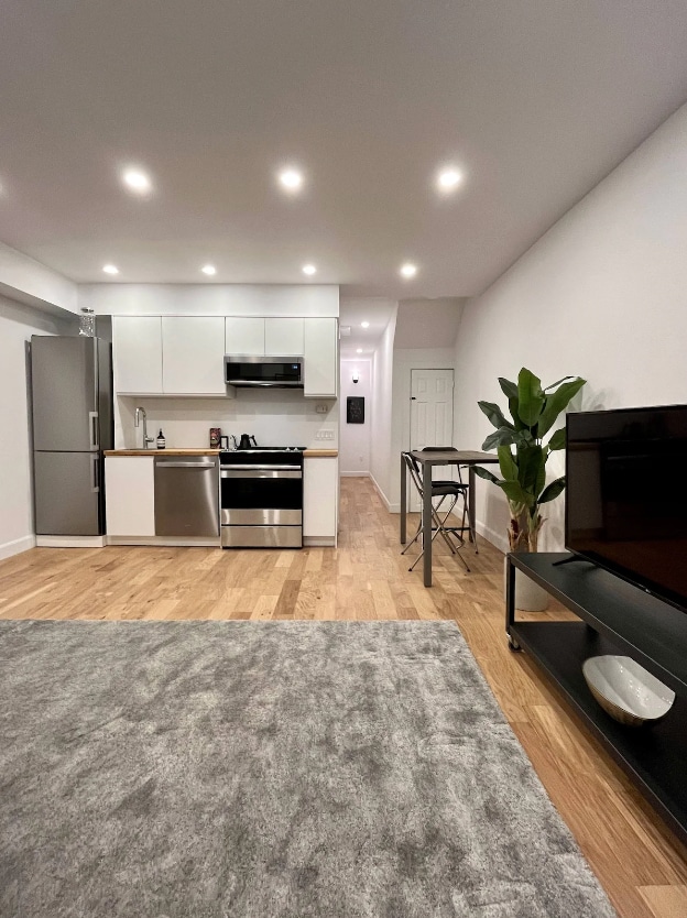 A modern kitchen features stainless steel appliances, including a refrigerator and oven. A sleek dining table for two is situated near a comfortable living area, which includes a TV on a minimalist stand. The space is illuminated by recessed lighting, enhancing the airy atmosphere.
