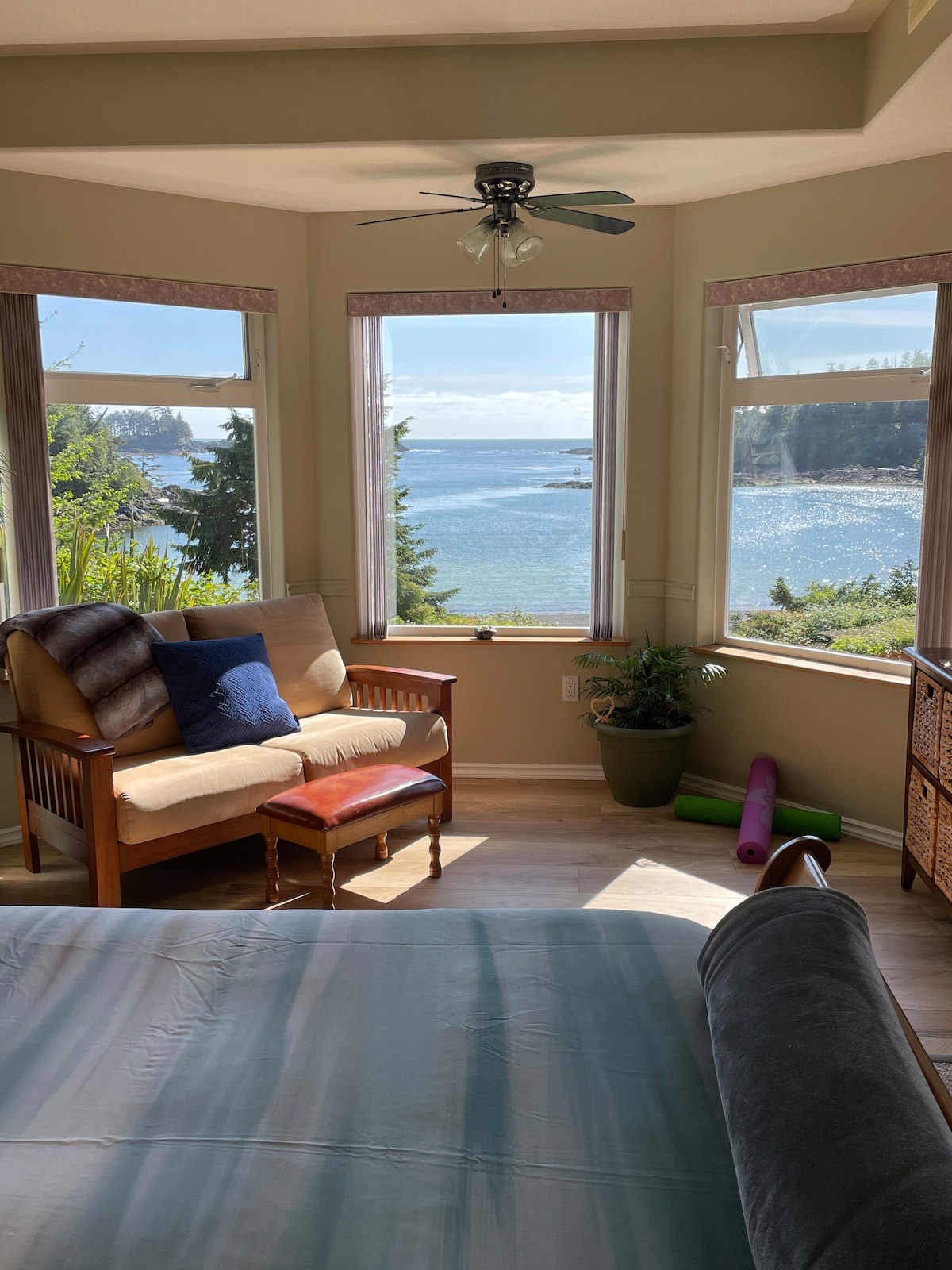 A cozy reading nook is positioned by expansive windows, providing unobstructed views of the ocean and surrounding trees. A wooden chair and footstool are complemented by a small side table. A yoga mat rests on the floor, enhancing the tranquil atmosphere.