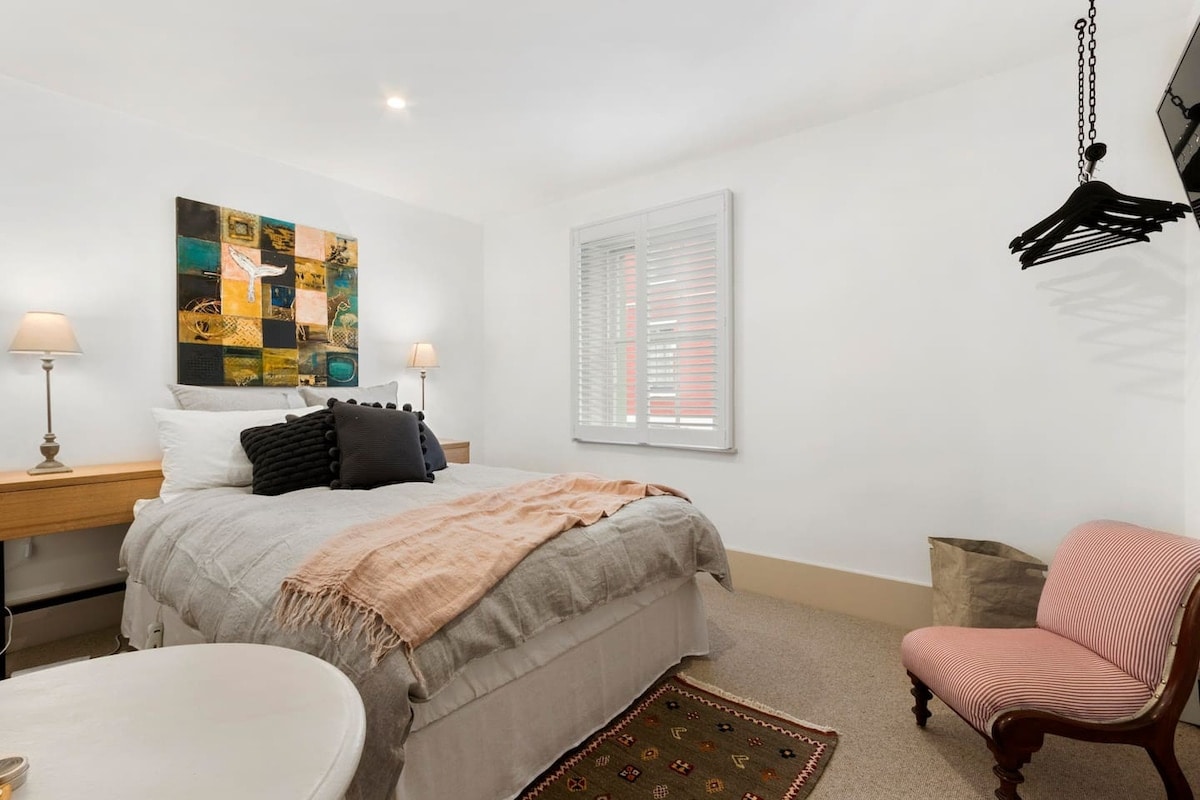 A cozy bedroom features a spacious bed, complemented by decorative pillows and a textured blanket. A colorful wall art piece brings character to the room. Soft lighting from bedside lamps enhances the inviting atmosphere. A small chair and hanging storage are positioned nearby.