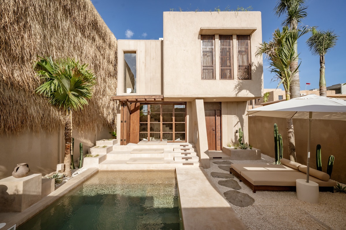 The image presents a contemporary retreat featuring a private pool surrounded by sleek stone steps. Tanned lounge chairs are arranged on a textured patio, complemented by tropical plants and cacti. The villa's earthy-colored walls and large windows offer a seamless connection to the outdoor space.