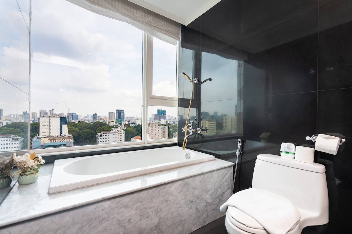 A modern bathroom features a freestanding bathtub situated beneath a large window, offering expansive views of the cityscape. The space is complemented by sleek black wall tiles and a marble countertop, with neatly arranged toiletries nearby.