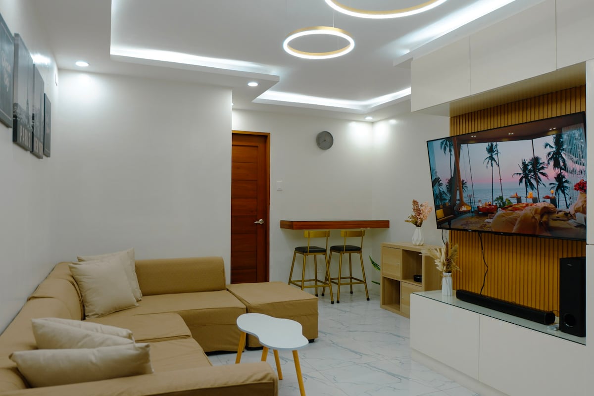 A modern living area is depicted, featuring a spacious beige sectional sofa adorned with soft pillows. A sleek white coffee table sits in front of the sofa. A large screen mounted on the wall displays a serene beach scene, while accent lighting illuminates the space.
