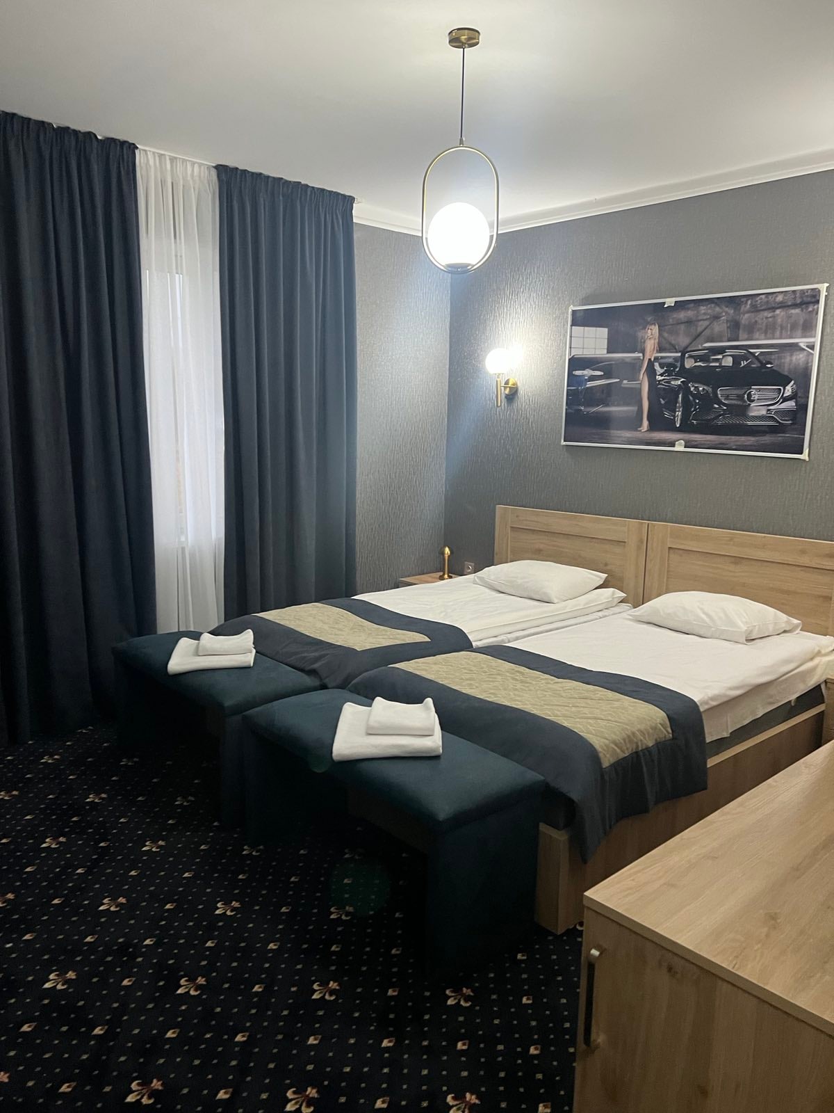 The sleeping area features two twin beds with crisp linens, topped with folded towels. A sleek wooden dresser is positioned nearby. Dark curtains frame the window, allowing natural light to filter in. A modern artwork piece hangs above the beds, adding a stylish touch.