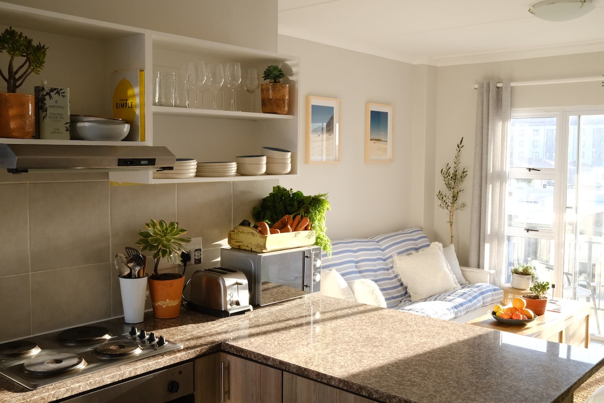 A compact kitchen area features a dark granite countertop with a stove and toaster, surrounded by open shelving displaying various kitchen items. A comfortable seating area with striped cushions is visible nearby, along with large windows that allow natural light to brighten the space.