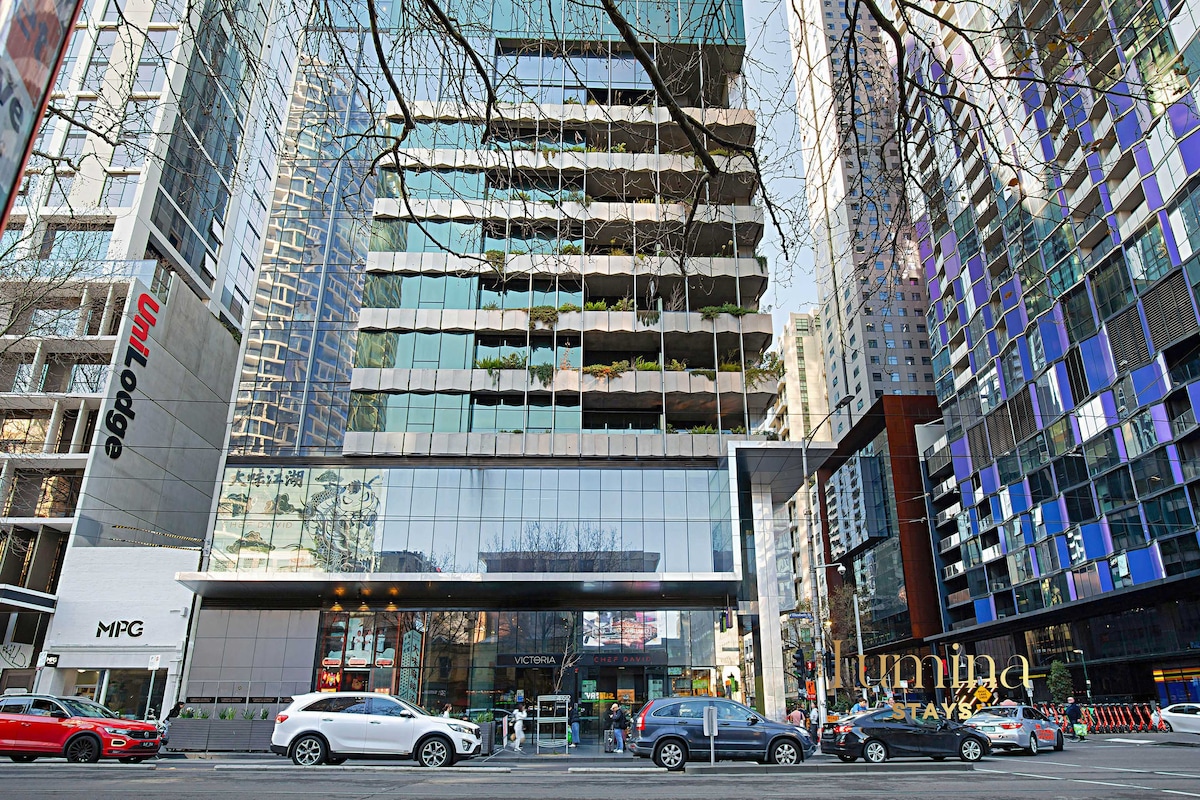 The modern multi-story building features a sleek glass facade reflecting the surrounding urban architecture. Greenery is visible on several balconies, and vehicles are seen parked along the bustling street in front, indicating a lively city atmosphere.
