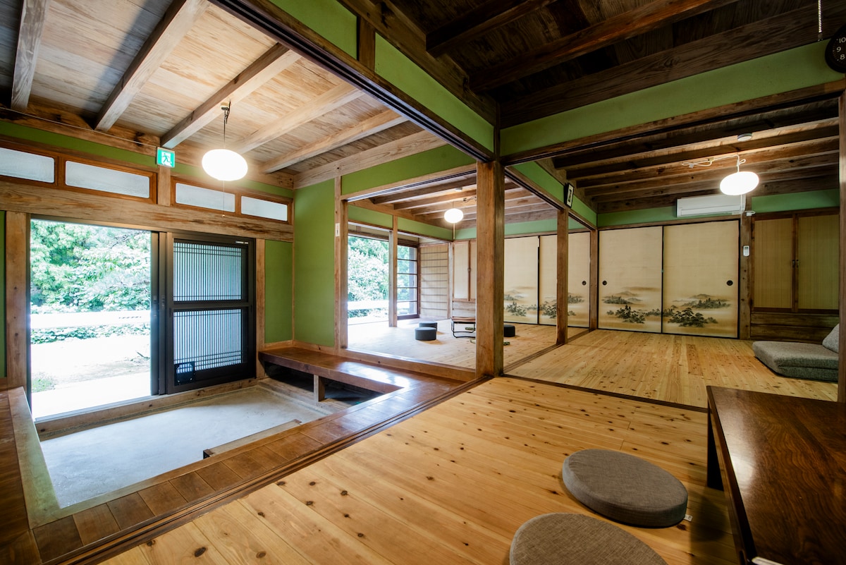 A spacious interior featuring natural wood flooring and exposed beams, with Shoji screens providing a seamless connection to the outdoor view. Low seating cushions are positioned around a wooden table, enhancing the layout for relaxation and social gatherings.