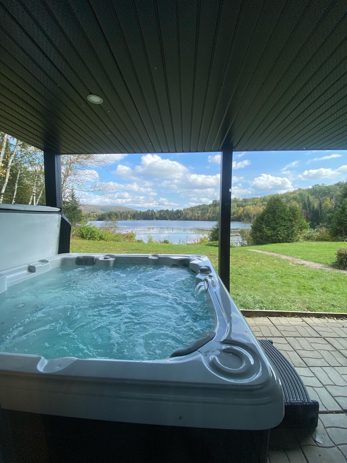 A hot tub is positioned on a private patio, surrounded by a covered structure. Beyond, a scenic view of the lake and lush greenery is visible, with clouds scattered across the sky. A pathway leads through the grass, enhancing the tranquil outdoor setting.