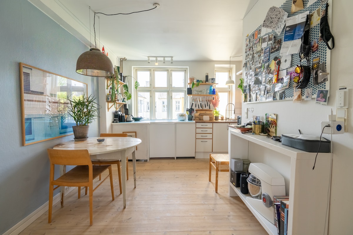 A light-filled kitchen area is presented with a round dining table and wooden chairs. A large window offers natural light, while an array of plants enhances the space. A wall covered with various items adds visual interest, alongside modern kitchen appliances.