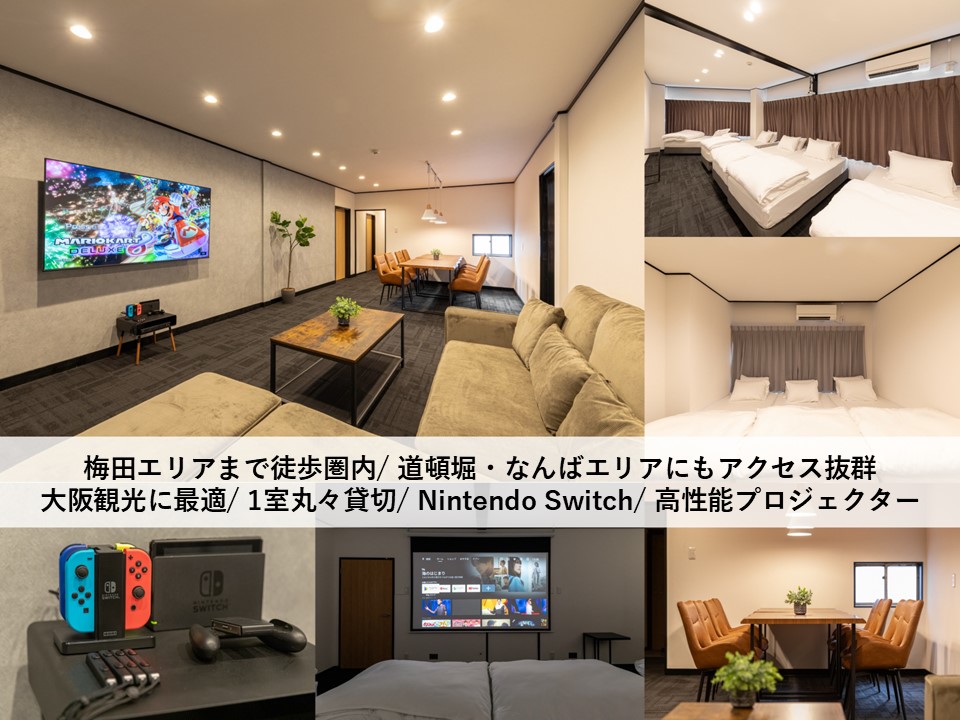 An inviting room features eight single beds with simple bedding. A large television is mounted on the wall, and a Nintendo Switch console is displayed on a table, alongside board games. Soft lighting enhances the relaxed atmosphere, while a projector is visible for shared entertainment.